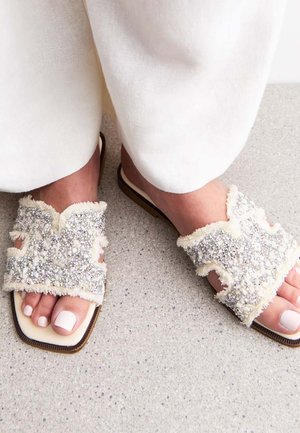 Sandali piatti con una tomaia in tessuto texturizzato beige, adornati con paillettes e perline argentate, presentano un design open-toe e un leggero tacco.