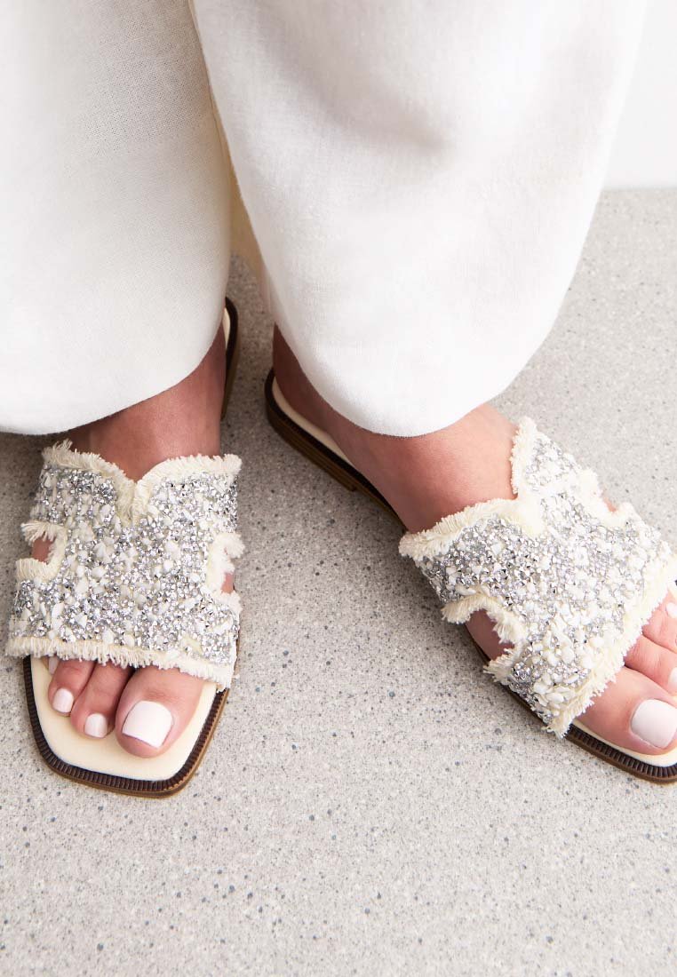 Platta sandaler med ett beige texturerat tygöverdrag, prydda med silverpaljetter och pärlor, med en öppen tå-design och en liten klack.