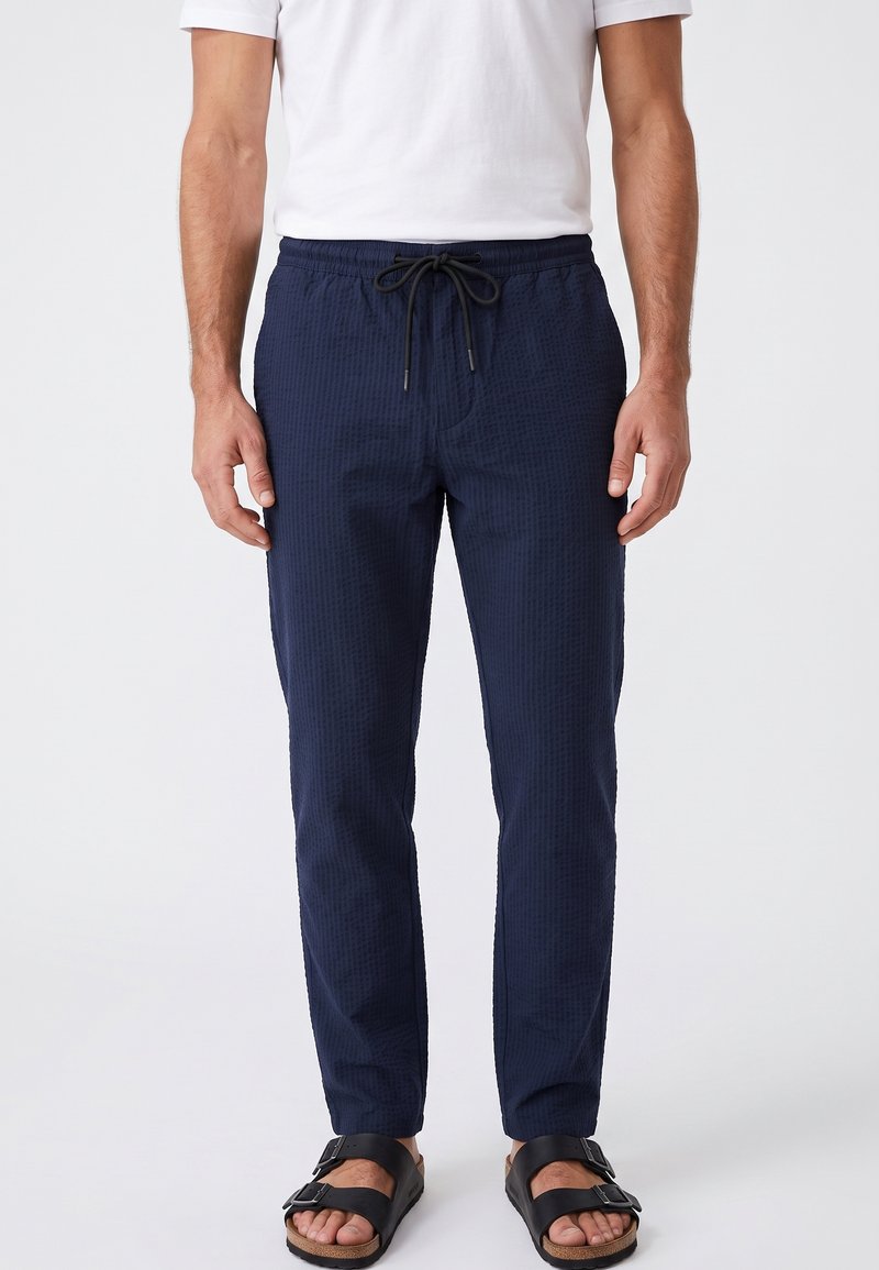 Hombre con pantalones azul marino con cordón, camisa blanca de manga corta y sandalias negras con hebilla, de pie frente a un fondo liso.