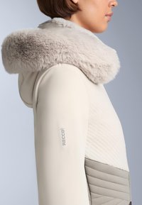 Világos bézs dzseki műszőrmés kapucnival, steppelt mintázattal és Recco márkajelzéssel az ujján. Sima tapintású és testhezálló kialakítású.
