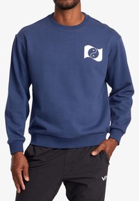 Sweat-shirt bleu marine en tissu doux, avec un logo abstrait blanc sur le côté gauche de la poitrine et des poignets et ourlet côtelés.