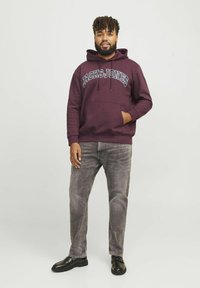 Bordeauxfarbener Hoodie mit "JACK & JONES"-Aufdruck auf der Brust, Fronttasche, kombiniert mit hellgrauen Jeans und schwarzen Schuhen, vor einem schlichten Hintergrund stehend.