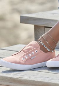 Pink canvas slip-on shoes with white rubber sole, featuring decorative side eyelet holes and a label. Ankle adorned with silver chain bracelets.