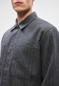 Szürke csíkos dzseki puha anyagból, két elülső zsebbel, gombos zárással és klasszikus galléros dizájnnal. Diszkrét logó részlet található rajta.