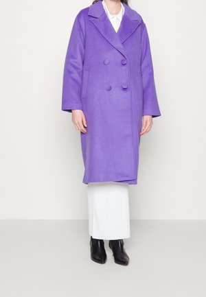 Osoba ubrana w długi fioletowy dwurzędowy płaszcz, białe spodnie i czarne botki, stojąca na tle jednolitego jasnego tła.