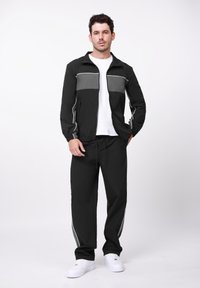 Μαύρο μπουφάν με γκρι πάνελ, φερμουάρ μπροστά και ελαστικά μανσέτες, σε συνδυασμό με μαύρο παντελόνι με λευκές πλευρικές ρίγες και λευκά αθλητικά παπούτσια.