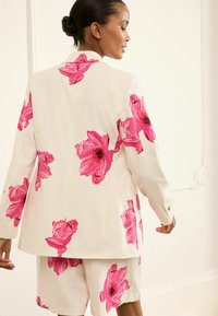 Weißes Blazer- und Shorts-Set mit großen rosa Blumendrucken. Der Stoff wirkt leicht und hat eine glatte Textur, akzentuiert durch goldene Knöpfe.