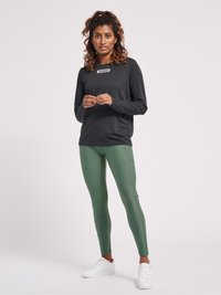 Zwart sporttop met lange mouwen en een logo op de borst, gecombineerd met strakke groene leggings en witte sneakers. Fijne, soepele stof.