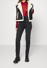 Chaleco de cuero sintético negro con cuello de borreguito crema, superpuesto a un suéter de cuello alto rojo, combinado con vaqueros negros de cintura alta y botas negras.