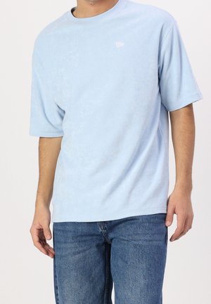 Homme portant un t-shirt uni bleu clair à manches courtes et un jean bleu, debout avec les bras détendus le long du corps devant un fond blanc.