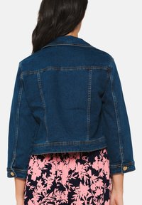 Blå denimjacka med kort skärning, knappdetaljer på ärmarna och synliga sömmar. Baksidan har en klassisk krage.