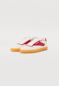 Hvite sneakers med røde skinndetaljer, teksturert overdel, kremfargede lisser og en gummisåle i en lys brunnyanse. Har detaljer med logo på siden.