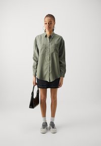 Olive green button-up shirt with two chest pockets, embellished with studs. Paired with a black denim skirt and gray sneakers.