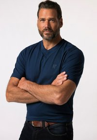 Camiseta de cuello en V azul marino hecha de tejido suave, con mangas cortas y un sutil logotipo en el pecho. El modelo posa con los brazos cruzados.