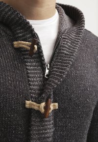 Grå stickad kofta med sjalkrage, med dragkedja och träkliknande detaljer fästa med jute. Strukturerad tygyta.