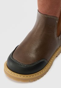 Close-up van een bruine leren laars met een zwarte neus, een tan rubberen zool en een bovenste gedeelte van bruin suède op een witte achtergrond.