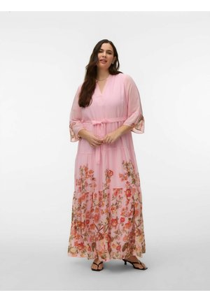 Ženska v dolgi svetlo roza obleki s cvetličnim vzorcem in zavezovanjem v pasu, stoji z rokami sklenjenimi, obuta v črne sandale z odprtimi prsti.