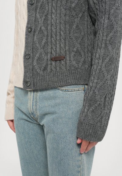 Grå kabelstickad cardigan med en krämfärgad sida, med runda knappar och en läderlogotyp, bärs över ljusblå denimjeans.