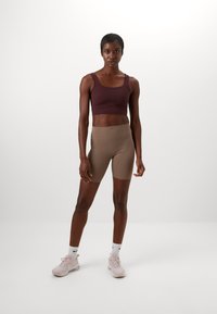 Bordó sportmelltartó széles pántokkal, barna testhezálló rövidnadrággal párosítva. A modellen rózsaszín sportcipők és fekete díszítésű fehér zoknik vannak.
