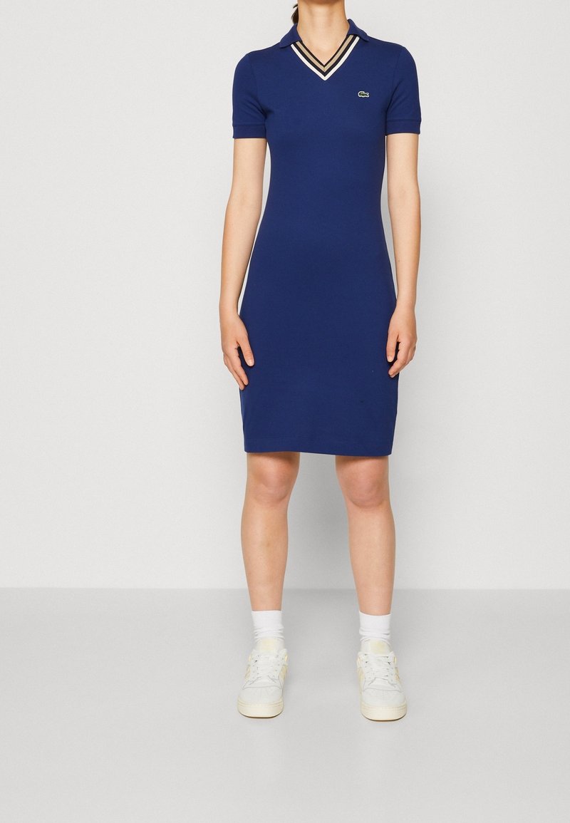Marineblaues Polokleid mit taillierter Passform, kurzen Ärmeln und V-Ausschnitt. Mit kontrastierendem gestreiften Besatz und kleinem gesticktem Logo.