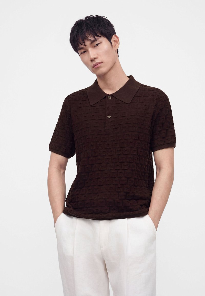 Joven con cabello corto y negro lleva una camiseta tipo polo de manga corta, texturizada y de color marrón oscuro, y pantalones blancos con las manos en los bolsillos.