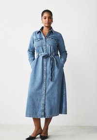 Denim-haalimekko, jossa on pitkät hihat, nappikiinnitys edessä, kaksi rintataskua, vyötärönauha ja maksi-pituus. Vaaleansininen väri ja pehmeä rakenne.