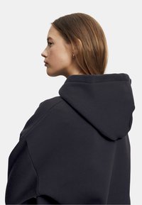 Mørkegrå hettegenser med raglanermer, myk tekstur og synlige sømdetaljer langs hetta og skuldersømmene.