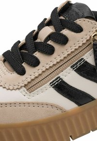 Beige sneaker featuring black laces and accents, a textured upper with suede and synthetic materials, and a ridged rubber sole.