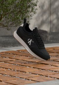 Μαύρο αθλητικό παπούτσι Calvin Klein με λευκή σόλα που αιωρείται πάνω από ξύλινο πλακόστρωτο πατάκι, με θολά φυλλώματα και πέτρινο τοίχο στο φόντο.