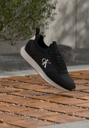 Svart Calvin Klein-sneaker med hvit såle som svever over en trebalkong, med uklart grøntområder og steinvegg i bakgrunnen.