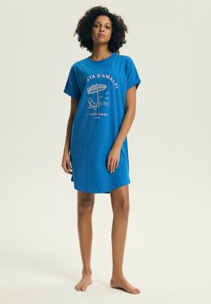 Kobieta stojąca boso, ubrana w niebieską sukienkę typu T-shirt z napisem "Costa d'Amalfi" oraz grafiką plażowego parasola i krzesła.