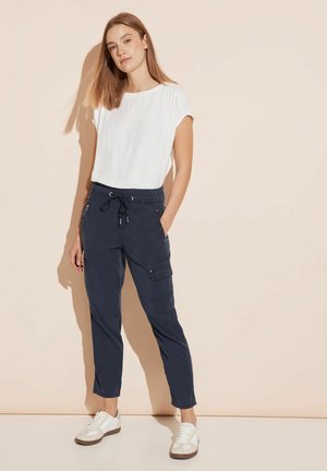 Weißes kurzärmliges Oberteil und marineblaue Cargo-Hose mit Taschen. Leichter Stoff, lockere Passform, kombiniert mit weißen Sneakers, stehend vor einem beigen Hintergrund.