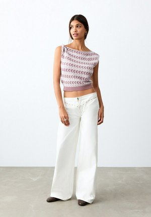 Ženska, oblečena v brezrokavnik s potiskom in široke bele hlače, stoji na betonski tla proti belemu zidu.