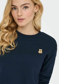 Sweatshirt bleu marine en coton, avec un petit motif de ours brodé sur la poitrine. Texture douce et col rond.