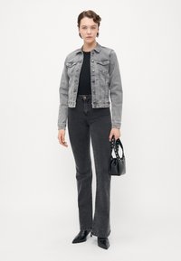 Kvinne iført en grå denimjakke, svart topp, svarte høyhalsbukser, svarte spisse støvler, og holder en svart håndveske med nagler.