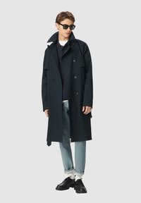 Uomo che indossa un trench nero doppiopetto, occhiali da sole neri, camicia scura, jeans chiari arrotolati, calzini bianchi e scarpe in pelle nere.