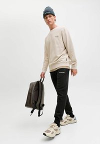 Beige sweatshirt met logo, zwarte sportbroek met zijstrepen, grijze beanie en bruine rugzak. Model draagt lichtgekleurde sneakers.