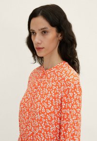 Chemise florale orange avec un motif de feuilles blanches, tissu doux, col rond et boutons à l'avant. Manches longues et coupe décontractée.