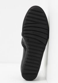 Semelle de chaussure noire avec des rainures horizontales et un logo, positionnée à la verticale contre un coin blanc avec des détails visibles du motif de la semelle.
