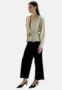 Blusa avvolgente beige con una cintura in vita, maniche lunghe e colletto, abbinata a pantaloni neri a gamba larga e tacchi a punta neri.