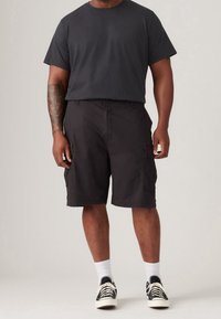 Black cotton t-shirt paired with black cargo shorts featuring side pockets, worn with white socks and black patterned sneakers.
