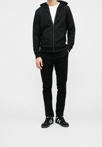 Černá mikina na zip s nastavitelnými šňůrkami, nošená přes bílé tričko. Kombinována s černými kalhotami a černými teniskami s bílými pruhy.