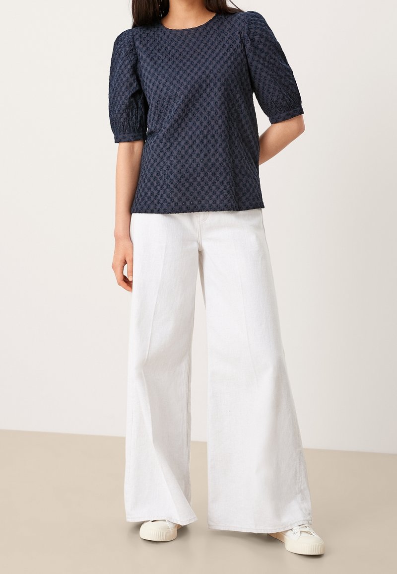 Top strutturato blu Navy con maniche a palloncino corte, abbinato a pantaloni bianchi a gamba larga e sneakers bianche. Il tessuto presenta un motivo a diamante.