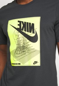 Grå bomullströja med en stor svart Nike-logga på en neongul grafik, med intrikata linjemönster och en mönstrad rutbas.