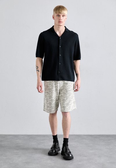 Schwarzes Kurzarmhemd mit Knöpfen, hell gemusterte Shorts, schwarze Socken und klobige schwarze Schuhe. Glatte Texturen und lässiger Stil.
