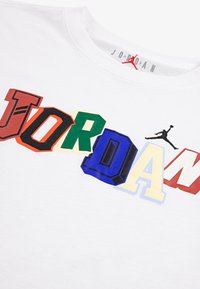 T-shirt bianco in cotone con lettere "JORDAN" sovrapposte in colori diversi, in grassetto e con font vari, e un logo Jumpman nero sul lato.