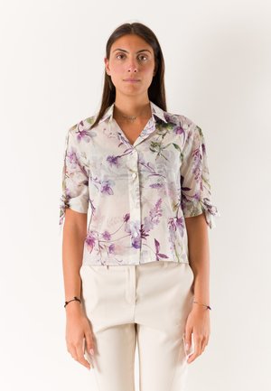 Donna che indossa una camicetta bianca trasparente con stampa floreale viola e maniche arrotolate, abbinata a pantaloni beige chiaro, in piedi davanti a uno sfondo semplice.