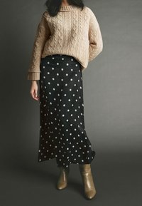 Beige kabelgebreide trui met verlaagde schouders, gecombineerd met een zwarte satijnen rok met witte stippen. Tan hakken enkellaarzen maken de look compleet.