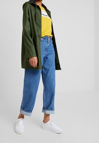 Groene waterdichte jas met zwarte capuchon, gele graphic t-shirt en hoogzakken blauwe spijkerbroek. Witte sneakers maken de outfit af.