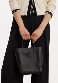 Melna teksturēta ādas rokassoma ar logo detaļu, kurai ir divas īsas rokas. To tur persona, kas ģērbusies bežā džemperī un melnās biksēs.